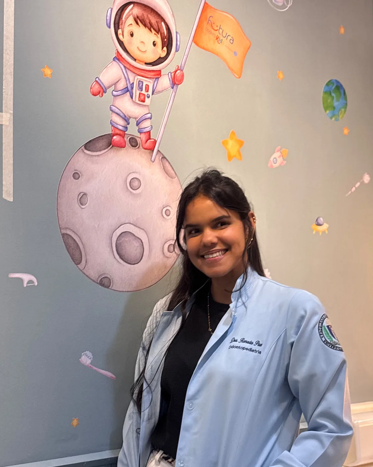Dra. Renata Paz no consultório de odontopediatria, ao lado da parede temática de astronauta